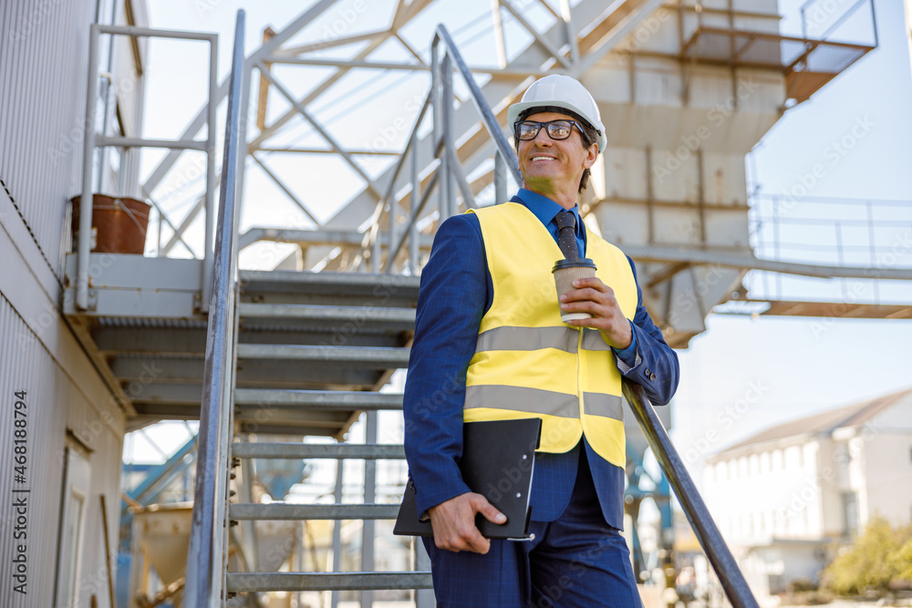 Cheerful man in work vest holding takeaway drink and folder while standing on staircase at manufacturing plant Cheerful man in work vest holding takeaway drink and folder while standing on staircase at manufacturing plant