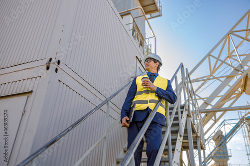 Smiling man factory worker holding takeaway drink and folder while standing on staircase outside industrial building