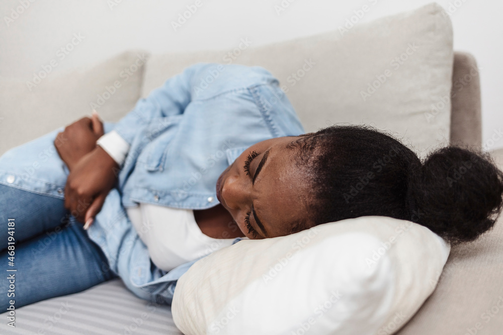 Cropped shot of an attractive young woman lying on her sofa alone and suffering from period cramps at home. Woman with menstrual pain. Woman in painful expression holding hands against belly. Cropped shot of an attractive young woman lying on her sofa alone and suffering from period cramps at home. Woman with menstrual pain. Woman in painful expression holding hands against belly.