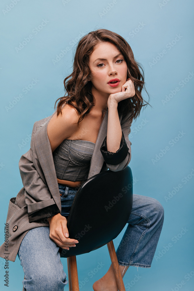 caucasian young woman sitting on high chair posing effectively against blue background. Sophisticated brunette with wavy hair is dressed in gray top, jacket top and jeans. caucasian young woman sitting on high chair posing effectively against blue background. Sophisticated brunette with wavy hair is dressed in gray top, jacket top and jeans.