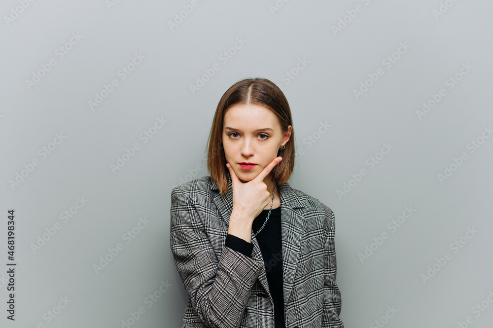 Portrait of a pensive lady in office clothes standing on a background of a gray wall and looking at the camera and thinking with a serious face, wearing a jacket. Portrait of a pensive lady in office clothes standing on a background of a gray wall and looking at the camera and thinking with a serious face, wearing a jacket.