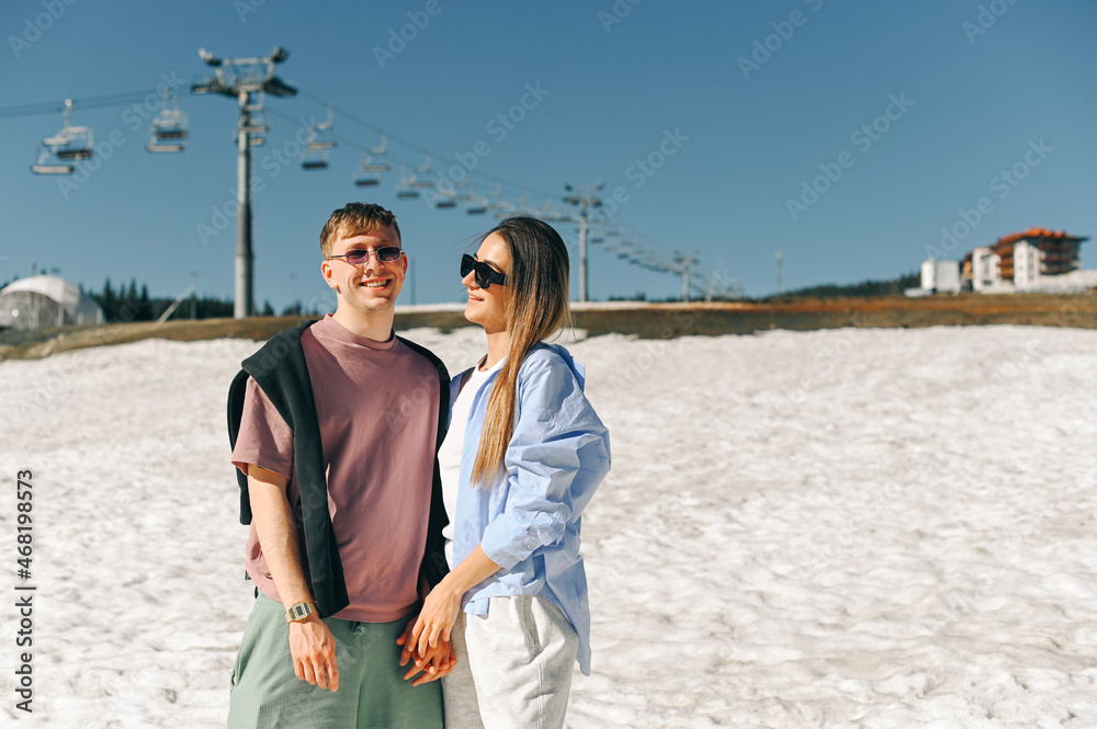 Young stylish couple in summer clothes standing on the snow in the mountains against the backdrop of ski lifts and hugging with a smile on their faces, posing for the camera