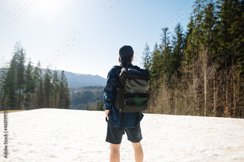 Man hiker in casual clothes and a cap with a backpack on his back stands on a snowy mountain and looks ahead, view of the back