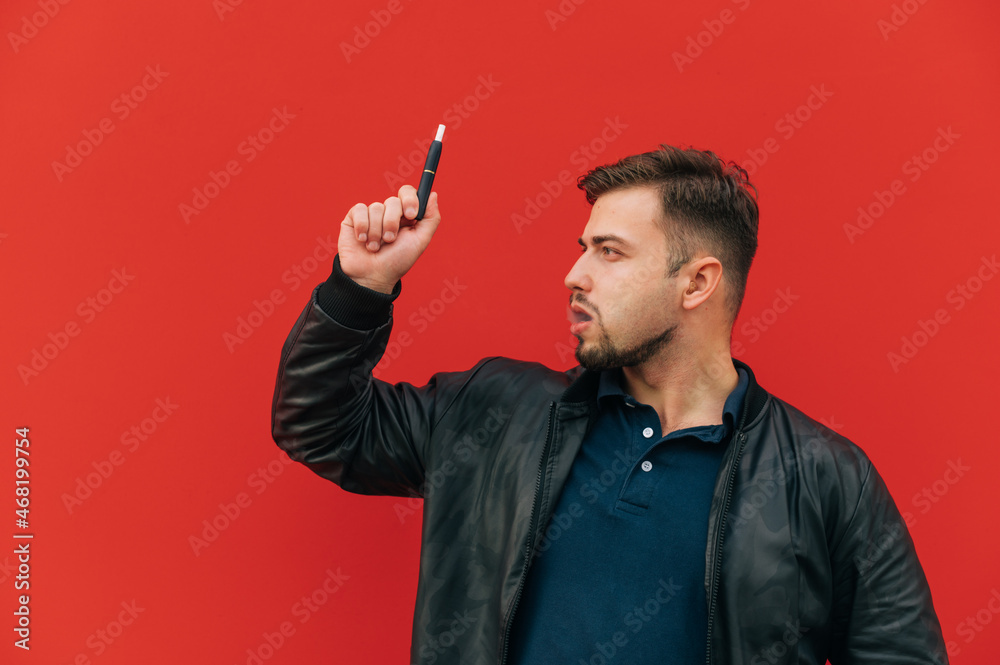 Portrait of handsome man with beard smokes e-cigarette heating tobacco with on red wall background and looks away with serious face. Portrait of handsome man with beard smokes e-cigarette heating tobacco with on red wall background and looks away with serious face.