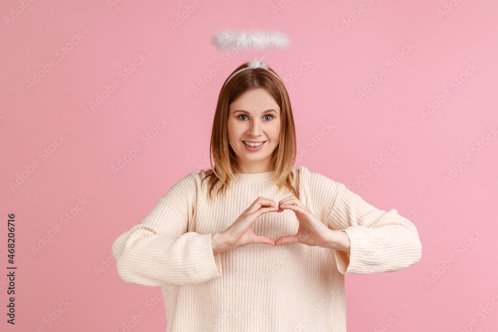 Portrait of cute pretty blond angelic woman with nimbus over head looking at camera and showing heart shape with hands, wearing white sweater. Indoor studio shot isolated on pink background.