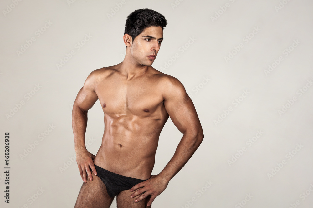 Fototapeta premium Portrait of handsome man with tanned athletic body, demonstrating his perfect sporty body, looking away, keeps hands on hips, wearing black panties. Indoor studio shot isolated on gray background.