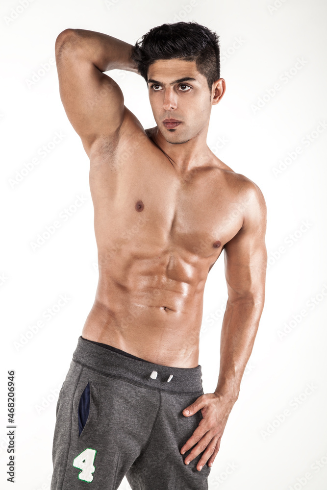 Obraz premium Portrait of young adult handsome brunette man posing shirtless with raised arm, looking away with serious expression, wearing trousers. Indoor studio shot isolated on white background.