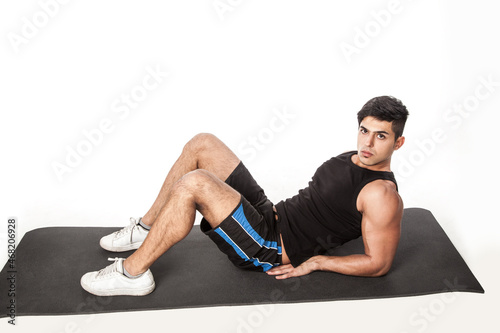 Portrait of handsome confident man lying on fitness mat, looking at camera, wearing sports clothing, doing abs exercises, healthy lifestyle. Indoor studio shot isolated on white background.