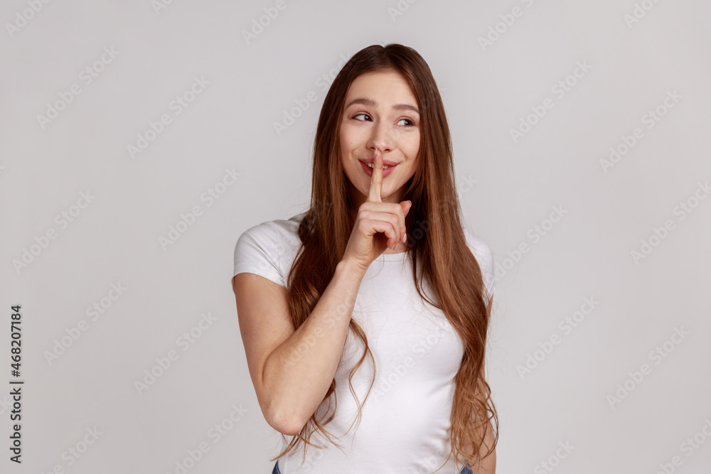 Shh, it's big secret. Beautiful cunning woman smiling, showing gesture secret sign with finger near her lips, wearing white T-shirt. Indoor studio shot isolated on gray background. Shh, it's big secret. Beautiful cunning woman smiling, showing gesture secret sign with finger near her lips, wearing white T-shirt. Indoor studio shot isolated on gray background.