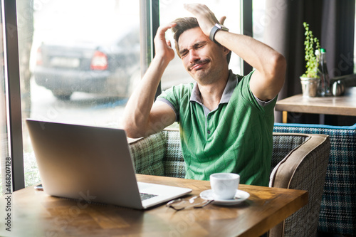 Canvastavla Portrait of desperate businessman wearing green T-shirt, feeling sorry and blaming himself for the mistake while working online on laptop, Indoor shot near big window, cafe background