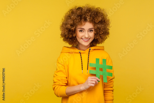 Happy beautiful woman holding green hashtag symbol, promoting viral topic in social network, tagging blog trends, wearing casual style hoodie. Indoor studio shot isolated on yellow background.