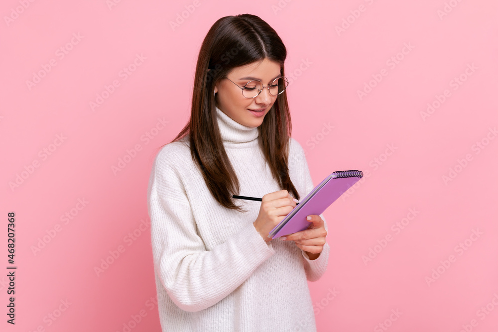 Smart positive female writing in paper notebook, making to do list or plan, outlines information, wearing white casual style sweater. Indoor studio shot isolated on pink background. Smart positive female writing in paper notebook, making to do list or plan, outlines information, wearing white casual style sweater. Indoor studio shot isolated on pink background.