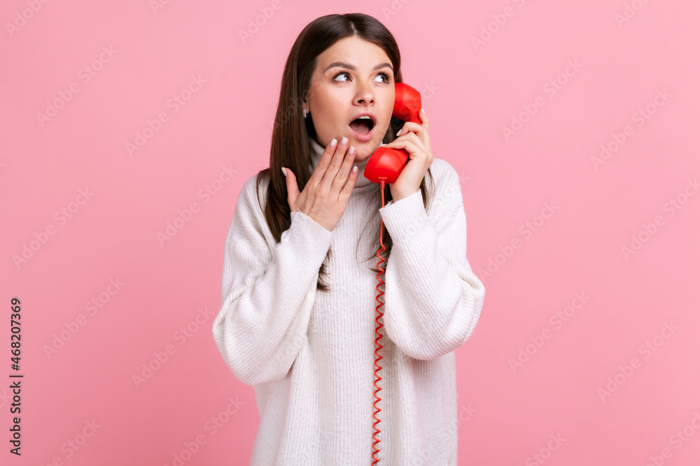 Portrait of shocked astonished woman talking red retro telephone, hearing shock news, open mouth, wearing white casual style sweater. Indoor studio shot isolated on pink background. Portrait of shocked astonished woman talking red retro telephone, hearing shock news, open mouth, wearing white casual style sweater. Indoor studio shot isolated on pink background.