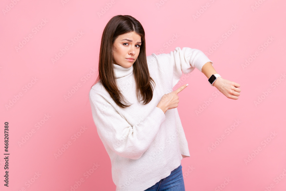 Portrait of impatient woman pointing wrist watch and looking displeased, showing clock to hurry up, wearing white casual style sweater. Indoor studio shot isolated on pink background. Portrait of impatient woman pointing wrist watch and looking displeased, showing clock to hurry up, wearing white casual style sweater. Indoor studio shot isolated on pink background.