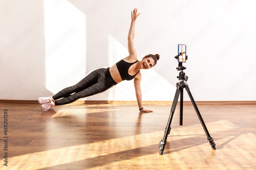 Side view of sporty woman recording video of doing side plank exercise, using phone camera on tripod, wearing black sports top and tights. Full length studio shot illuminated by sunlight from window. Side view of sporty woman recording video of doing side plank exercise, using phone camera on tripod, wearing black sports top and tights. Full length studio shot illuminated by sunlight from window.