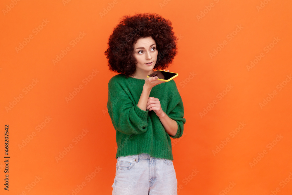 Digital speaker app. Portrait of woman with Afro hairstyle in green sweater talking to mobile phone, using voice assistant to record reminder, message. Indoor studio shot isolated on orange background