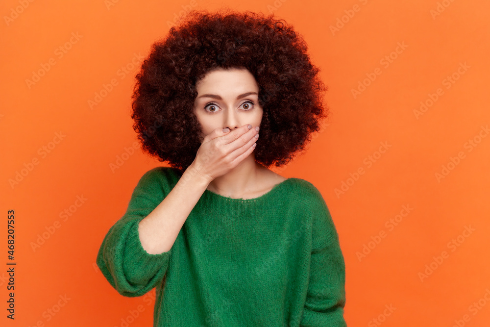 Young adult woman with Afro hairstyle wearing green casual style sweater covering mouth with palm, keep secret, does not tell anyone. Indoor studio shot isolated on orange background. Young adult woman with Afro hairstyle wearing green casual style sweater covering mouth with palm, keep secret, does not tell anyone. Indoor studio shot isolated on orange background.