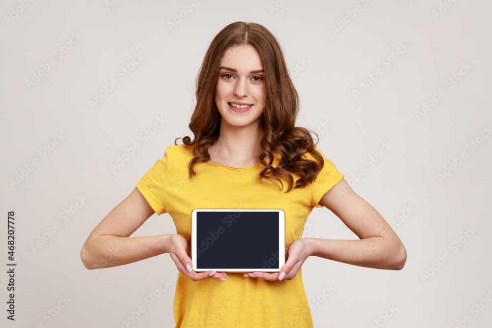 Attractive woman of young age holding and showing tablet with blank screen with toothy smiling and satisfied face, copy space for advertisement. Indoor studio shot isolated on gray background. Attractive woman of young age holding and showing tablet with blank screen with toothy smiling and satisfied face, copy space for advertisement. Indoor studio shot isolated on gray background.