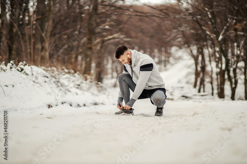 Fit sportsman kneeling on snowy path in nature and tying shoelace. Winter fitness, outdoors sport, sportswear