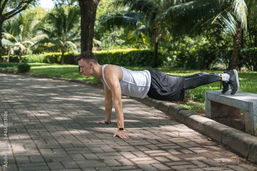 Sporty active young man in sportswear doing sport exercises in the park. Fit caucasian man doing push ups in the park on the bench. Healthy lifestyle
