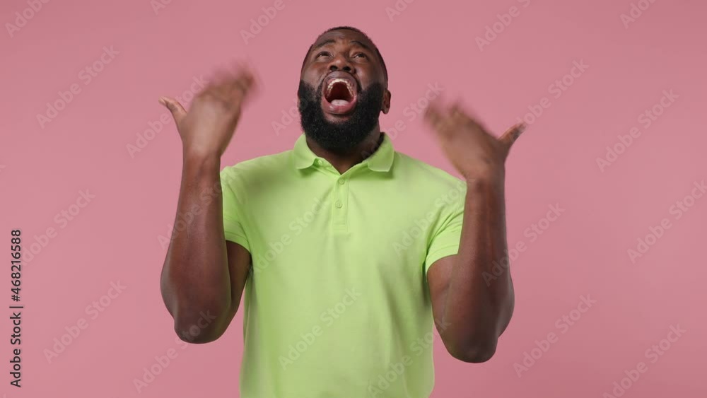 Young african american man 20s wears green t-shirt after black film making clapperboard play act cry shout scream why look up raise hands isolated on plain pastel light pink background studio portrait