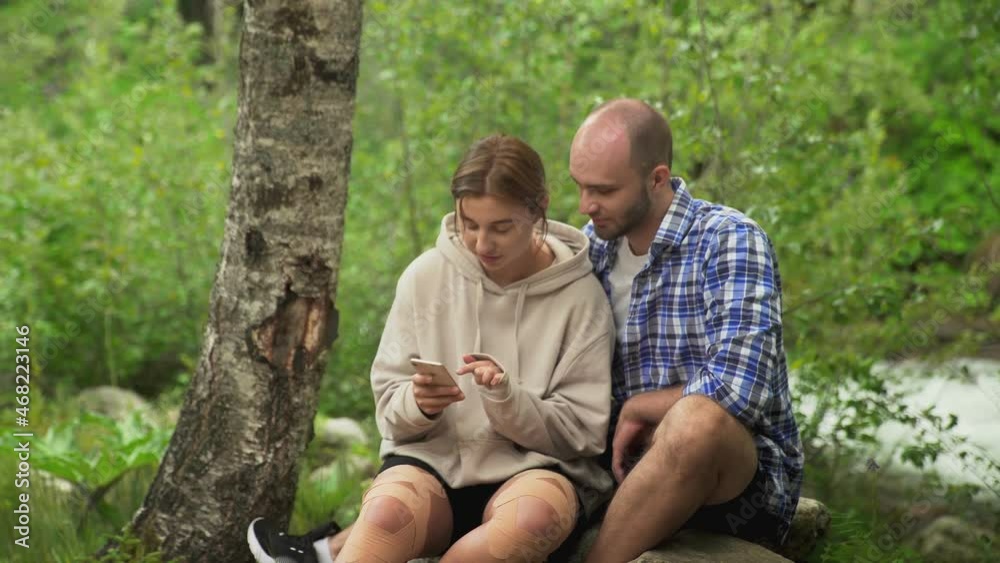 A man and a woman are sitting on a rock. A woman shows a man something on her phone. A boy and a girl have a date. In the forest outdoors A man and a woman are sitting on a rock. A woman shows a man something on her phone. A boy and a girl have a date. In the forest outdoors