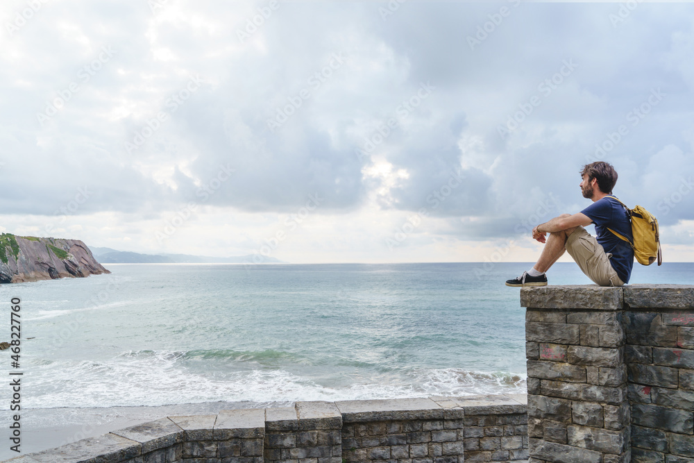 Panoramic view of young traveler man sit in a cliff. Horizontal view of backpacker traveling outdoors with the blue ocean on the background.