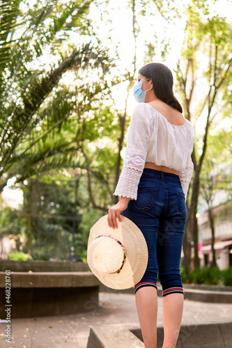young latin tourist woman, standing in a park enjoying the afternoon, with a hat in her left hand, wearing a protective mask, model not recognizable.