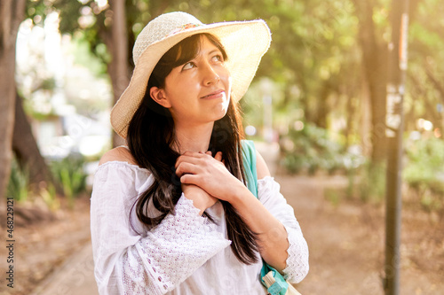 young latin woman, hands on chest, smiling face looking up, beam of light, fleer, white blouse and trees in background. photo
