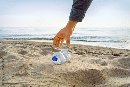 An arm reaches to pick up an empty bottle on a beach