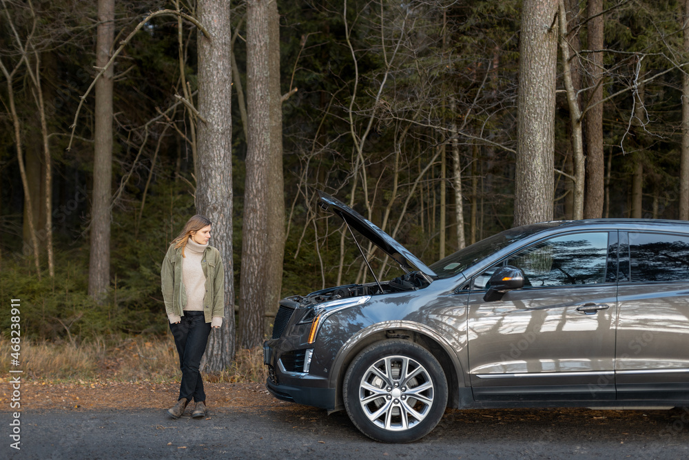 Sad Caucasian girl stands at the open hood of car. Automobile broke down on country road. Woman need auto mechanic for broken car. Copy space. Side view. Sad Caucasian girl stands at the open hood of car. Automobile broke down on country road. Woman need auto mechanic for broken car. Copy space. Side view.