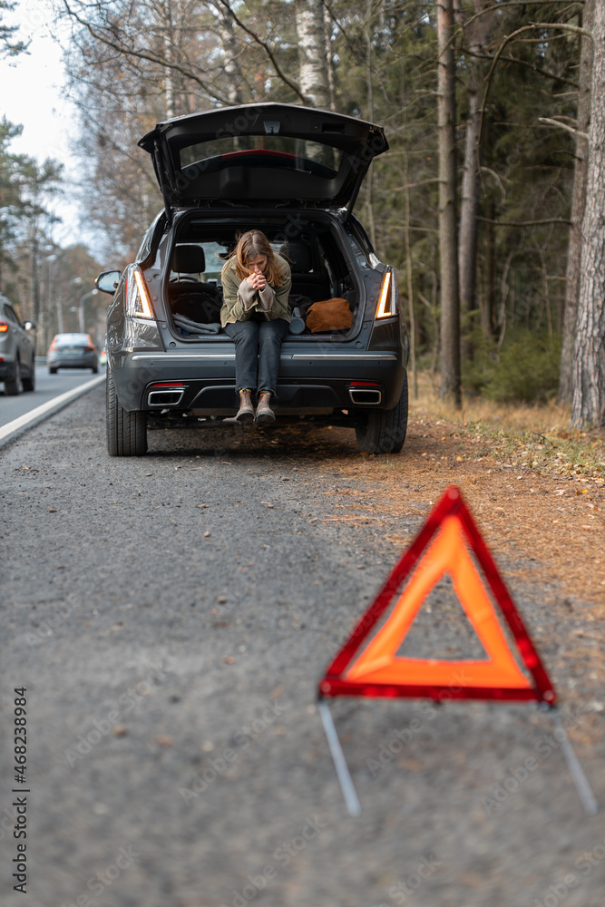 Sad Caucasian girl sitting in trunk of broke down car. On the foreground emergency stop sign. Selective focus.