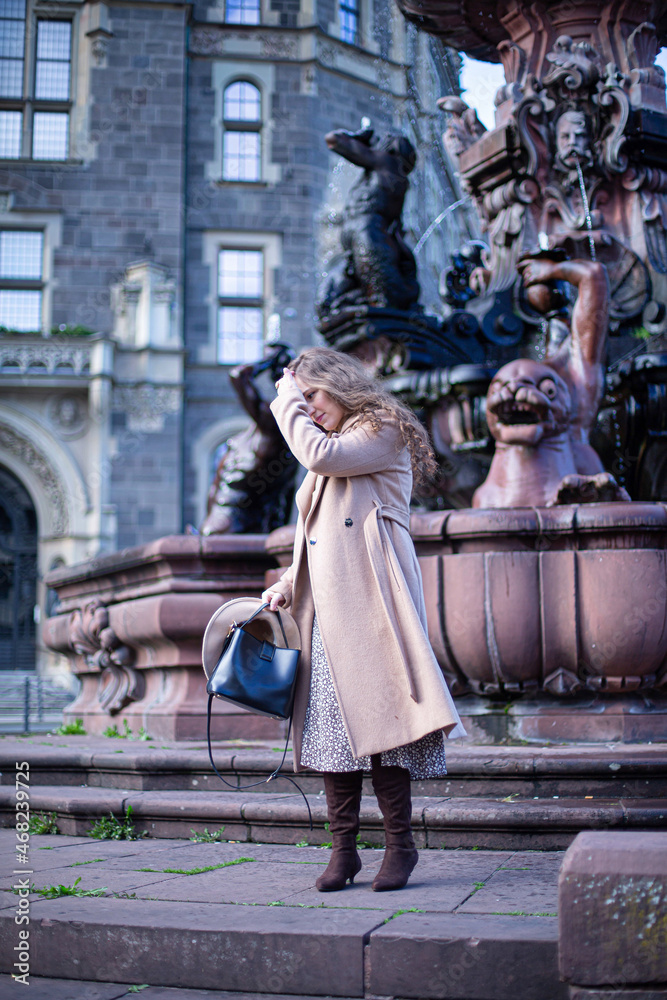 Pretty woman with curly hair in autumn coat with fountain behind holding hand near her face