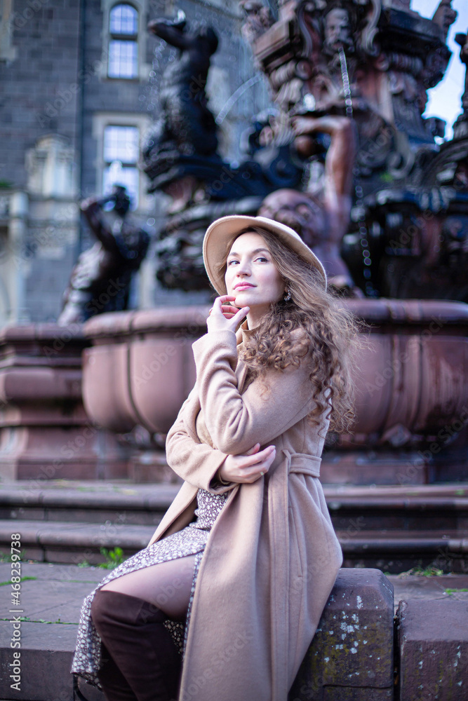 Pretty woman with curly hair in autumn coat with fountain behind holding hand near her chin