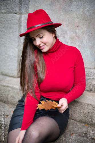 Portrait of white woman with long dark hair in bright red near stone wall holding autumn leaves in her hand © PIXbank