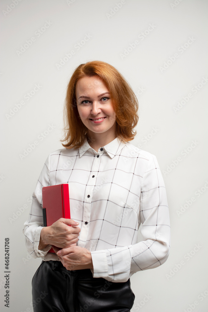 a girl with golden hair in a white blouse and black leather trousers holds a red notebook in her hands. white background.