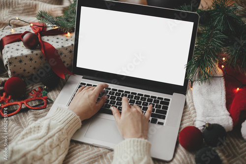Hands working on laptop with blank screen on cozy bed with stylish christmas ornaments, present and santa hat. Christmas shopping online and freelance in holidays. Space for text © sonyachny