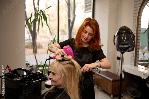 Fashionable beautiful red-haired female hairdresser using a round comb and hair dryer straightens blonde hair to a client of a beauty salon, performing her a stylish hairstyle. Beauty industry concept