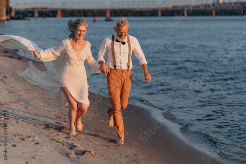 Cheerful elderly couple runs along beach holding hands. © MZaitsev