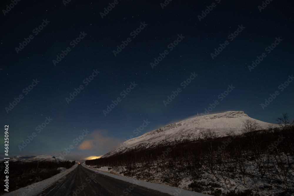 Naklejka premium Borealis aurora sweeps over a main road on a cloudless night in Kilpisjarvi, Lapland, Finland. aurora polaris in green dances across the sky. Scandinavian magic. Saana mountain