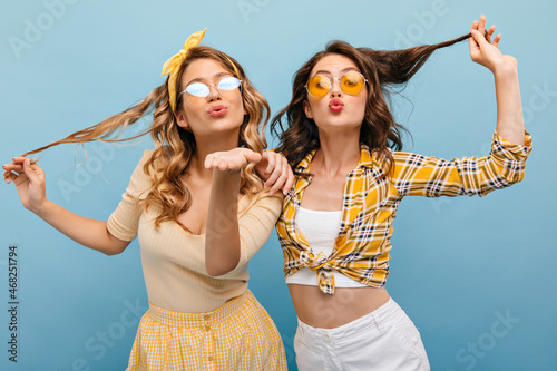 Two happy cute caucasian young girls on blue isolated background send air kiss to camera. Beauties with wavy hair wearing glasses are dressed in light casual clothes. 