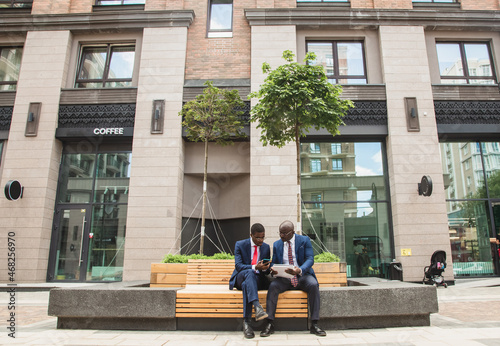 Two dark-skinned african american businessmen in suits with briefcases sit on a bench against the backdrop of a building facade
