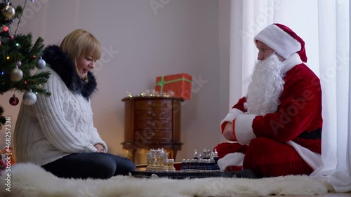 Smiling adult woman and man in Father Christmas costume playing chess sitting on soft carpet in living room at home, Cheerful Caucasian husband and wife enjoying New Year together indoors