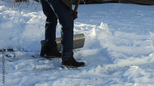 Person is snow shovelling on a winter day. Man is wearing jeans and winter boots, using a snow shovel to remove snow from the driveway.