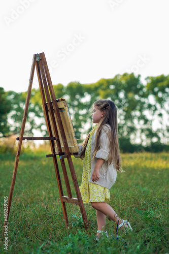 A little girl stands in a field during the day and paints a picture on an easel