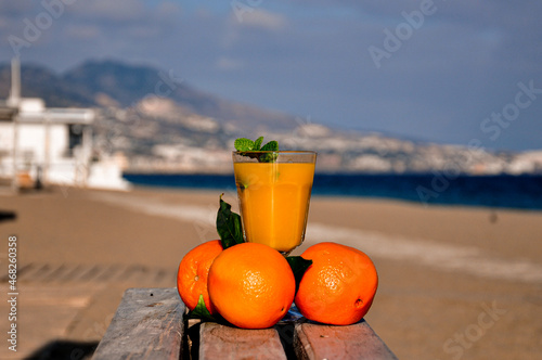 A glass with orange juice and a sprig of mint surrounded by three oranges is on a wooden table against a blurred background of the beach, sea, mountains and resort town in clear weather.