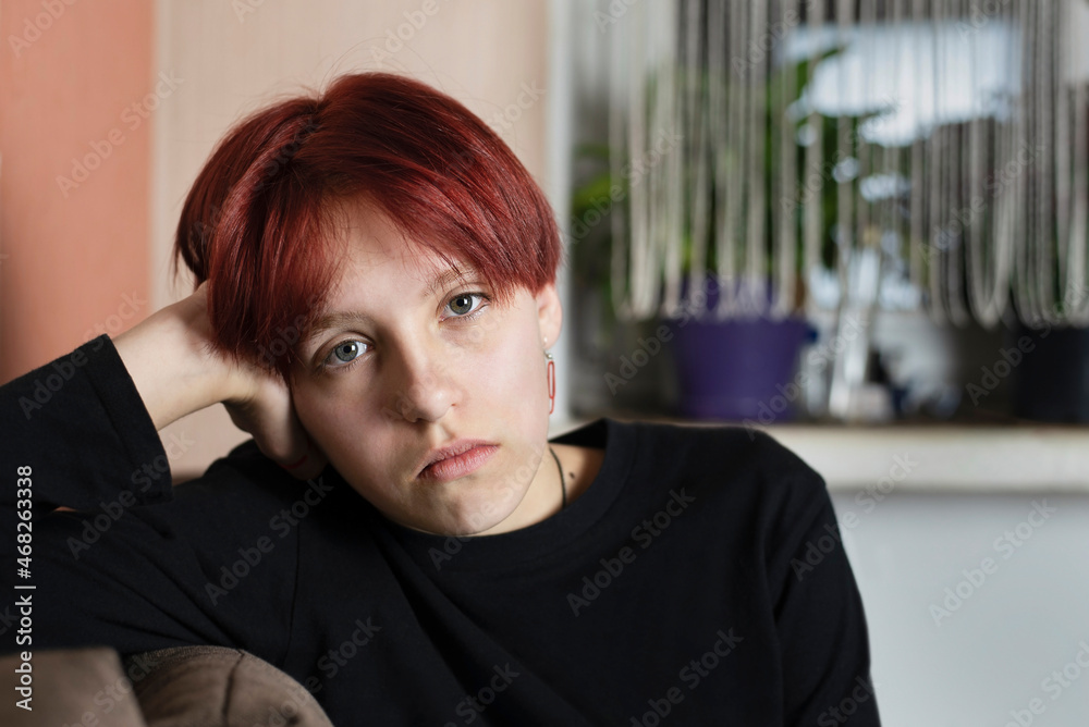 Fototapeta premium A teenage girl with red hair and complex character and a sad face is sitting at home, she quarreled with her friends and has no one to be friends with, she feels lonely