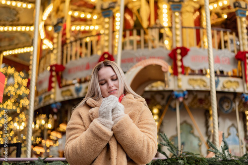 Cozy winter portrait of glorious young woman  standing near christmas carousel.  Model wearing warm white woolen gloves and warm coat