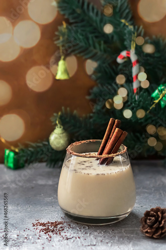 Christmas eggnog with cinnamon sticks on a background of fir branches decorated with toys, blurred bokeh. Glass of a festive winter drink on a concrete background with copy space. 