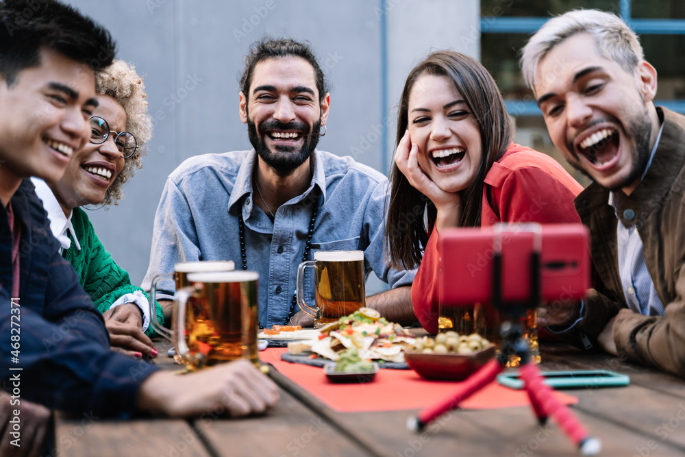 Obraz premium Group of multiracial friends enjoying food and drinking beer while making online video call with mobile phone at brewery bar to celebrate birthday - Focus on bearded man face